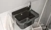 A dark-colored sink with a chrome faucet is mounted on a tiled wall. A white towel hangs to the left, and a mop bucket with a mop is to the right. Náhled obrázku produktu
