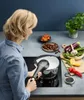 A woman is using a pressure cooker in a kitchen setting, with various ingredients like vegetables and dates laid out on the counter. Náhled obrázku produktu