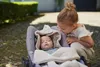 A young girl looks down at a baby sleeping in a car seat outdoors. Anteprima dell’immagine prodotto
