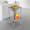 A stainless steel kitchen sink with a faucet, filled with various fruits, is placed on a stand in a kitchen setting. A large orange bottle is on the shelf below. Náhled obrázku produktu