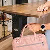 A person is holding a pink bag under a brown wooden desk. Produktbild-Vorschau