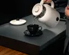 A person is pouring hot water from a white electric kettle into a black teacup and saucer. Podgląd zdjęcia produktu