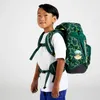 A young boy wearing a white t-shirt and blue jeans stands against a white background, modeling a green backpack with a yellow geometric pattern. Produktbild-Vorschau