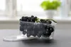 A bunch of black grapes in a clear plastic container with its lid beside it, placed on a dark gray surface. The background is blurred, showing a plant and a window. Produktbild-Vorschau