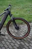 A close-up of the front wheel and frame of a gray and black electric mountain bike with tan tires, resting on a paved walkway with grass in the background. Produktbild-Vorschau