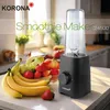 A Korona Smoothie Maker is displayed on a kitchen counter next to a bowl of fresh fruits, including bananas, strawberries, and apples. The background shows a kitchen setting with sunlight streaming through a window. Produktbild-Vorschau
