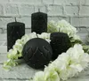 Four black candles of different shapes and sizes are arranged with white flowers against a white brick wall. Produktbild-Vorschau