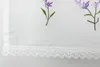 A close-up of a white fabric with embroidered lavender flowers and a lace trim. Produktbild-Vorschau