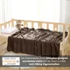 A brown fleece blanket is draped over a wooden crib, with a pillow and a teddy bear on the crib. There is a marketing overlay at the bottom with text and a logo. Produktbild-Vorschau