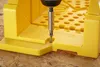 A close-up shot of a yellow plastic miter box with a screw being driven into it by a power drill. Náhľad obrázku produktu