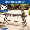 A metal garden bench with ornate scrollwork is shown outdoors on a paved patio surrounded by plants and flowers. Anteprima dell’immagine prodotto
