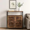 A dark brown wooden cabinet with slatted doors and a black metal frame, placed against a white wall. A framed picture and a vase with greenery are on the shelf above the cabinet. A portion of a gray sofa is visible to the right. Produktbild-Vorschau