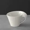 A white ceramic coffee cup with a unique handle sits on a gray surface against a black background. Produktbild-Vorschau