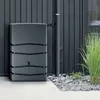 A gray rectangular water butt is positioned against a dark gray vertical slatted wall, next to a downpipe. The butt has a tap on the front. It is surrounded by light gray paving stones and gravel, with some green grass to the right. Anteprima dell’immagine prodotto