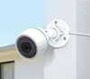 A white security camera mounted on a white wall, with a blurred background of a house and sky. Voir les images du produit