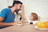 A father and child are at a table, with the child feeding the father. Voir les images du produit