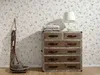 A dresser with drawers and a lamp on top, next to a decorative wooden object and baskets, against a floral wallpaper. Produktbild-Vorschau