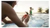 Two women on a surfboard in the water, one holding a small orange camera. Anteprima dell’immagine prodotto