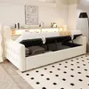 A beige upholstered bed with its storage compartment open, revealing folded bedding and pillows. The bed is set in a room with a patterned rug and a shelf with decorative items and lighting. Produktbild-Vorschau