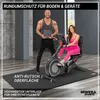 A man and a woman are in a home gym setting. The woman is using an exercise bike, and the man is standing next to her holding a tablet. They are both on a large black mat. Produktbild-Vorschau