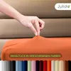 A hand is shown stretching an orange fitted sheet over a mattress. Below the sheet, a row of different colored swatches is displayed. Produktbild-Vorschau