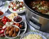 A slow cooker filled with food, surrounded by various dishes of food, including a salad, meatballs, olives, and rice. Náhled obrázku produktu