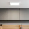 A square white LED ceiling light is mounted on a white ceiling above a modern kitchen with gray cabinets and a wooden countertop. Náhľad obrázku produktu