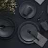 A dark, flat surface with a set of dark gray stoneware dishes, mugs, and cutlery, along with a small bunch of dried flowers. Náhľad obrázku produktu