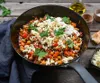 A close-up overhead shot of a skillet filled with a hearty meal, topped with feta cheese and parsley, set on a rustic wooden table. Náhled obrázku produktu