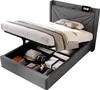 A grey upholstered bed with its storage compartment open, revealing folded clothes and storage boxes. Produktbild-Vorschau
