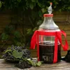 A large glass carboy with a fermentation lock, filled with dark liquid, sits on a wooden table next to a bowl of dark berries and some leaves. A red ribbon is tied around the carboy. Produktbild-Vorschau