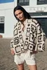 A woman stands outdoors in front of a white building, wearing a crocheted cardigan and shorts. Produktbild-Vorschau