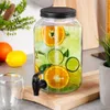 A glass beverage dispenser filled with water, ice, and slices of oranges and limes, placed on a wooden board on a countertop. There are also fruits and a glass in the foreground and a plant in a pot in the background. Produktbild-Vorschau