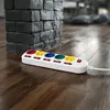 A white power strip with multiple colored outlets and individual switches rests on a polished wooden floor, next to a dark brick column. Produktbild-Vorschau