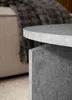 Close-up of a modern round coffee table with a textured gray surface and a geometric base, placed in a living room setting with a blurred sofa and throw blanket in the background. Voir les images du produit