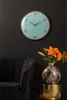 A wall clock is displayed on a dark wall above a black chair and a vase of orange flowers on a gold stand. Produktbild-Vorschau
