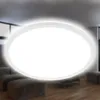 A modern, round LED ceiling light with a slim profile, emitting a bright, neutral white light and a subtle backlight effect, installed in a living room setting with blurred furniture in the background. Náhľad obrázku produktu