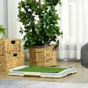 A pet toilet with artificial grass is placed on a wooden floor, next to woven baskets and a large potted plant. Produktbild-Vorschau