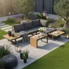 A modern outdoor lounge set with a sectional sofa, coffee tables, and decorative plants, set on a paved patio with grass and trees in the background. Produktbild-Vorschau
