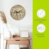 A wooden wall clock with a botanical design is displayed on a white wall above a wooden console table. To the right, there are two circular icons with text indicating sustainable wood and light weight. Produktbild-Vorschau