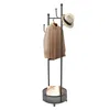 A black coat rack with a basket at the bottom, holding a beige handbag. A brown shirt is hanging on one of the hooks, and a beige hat is on another hook. Podgląd zdjęcia produktu