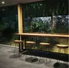 A bar counter with stools in front of a large window overlooking a lush green garden. Produktbild-Vorschau