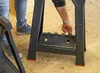 A person's hand is shown adjusting a black multi-functional stand, with another stand and people's legs visible in the background. Produktbild-Vorschau