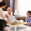 A patient in a hospital bed is smiling and interacting with a visitor and a nurse. Produktbild-Vorschau