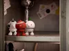 Three small, stylized figurines resembling sports balls with legs and faces are arranged on a shelf inside a locker. Produktbild-Vorschau
