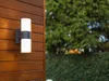 An outdoor wall light mounted on a wooden wall, with greenery in the background. Podgląd zdjęcia produktu