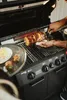 A person is using a rotisserie attachment on a grill to cook meat, with some food on a plate nearby. Produktbild-Vorschau