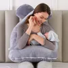 A woman is holding a baby while sitting on a bed, using a large U-shaped pillow. Náhled obrázku produktu