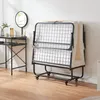 A folded metal rollaway bed with a mattress is leaning against a white wall, next to a black vanity table with a mirror and toiletries. Produktbild-Vorschau