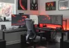 A gaming desk setup with multiple monitors, a chair, and ambient red lighting. Produktbild-Vorschau